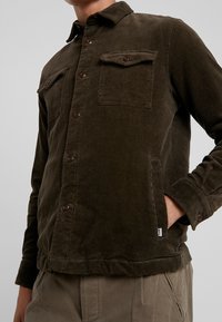 Veste-chemise en velours côtelé marron avec deux poches poitrine, fermeture à boutons, et boutons marron. Texture douce avec motif côtelé visible.