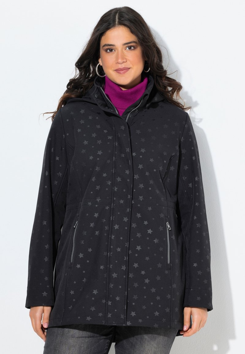 Žena oblečená v černé bundě s hvězdicovým vzorem na zip přes fialový rolák, stojí proti bílému pozadí, čelí dopředu.