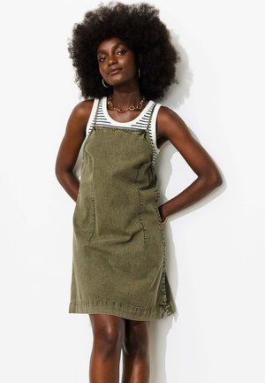 Femme avec une grande coiffure afro portant une robe salopette vert olive sur un débardeur rayé noir et blanc, debout les mains dans le dos.