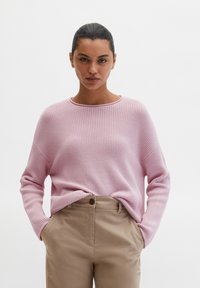 Femme aux cheveux foncés portant un pull en maille rose clair et un pantalon beige, debout les mains dans les poches devant un fond blanc uni.