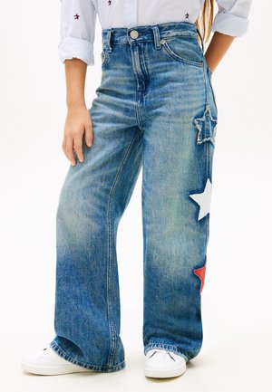 Barn med vide blå jeans med røde, hvite og frynsete stjernepatcher på siden, sammen med hvite joggesko og en hvit skjorte.