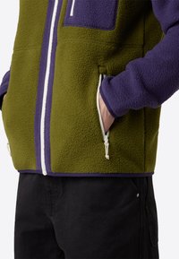 Giacca in fleece di colore verde oliva e viola con dettagli in bianco nella cerniera e due tasche laterali con zip, realizzata in un tessuto testurizzato e accogliente.