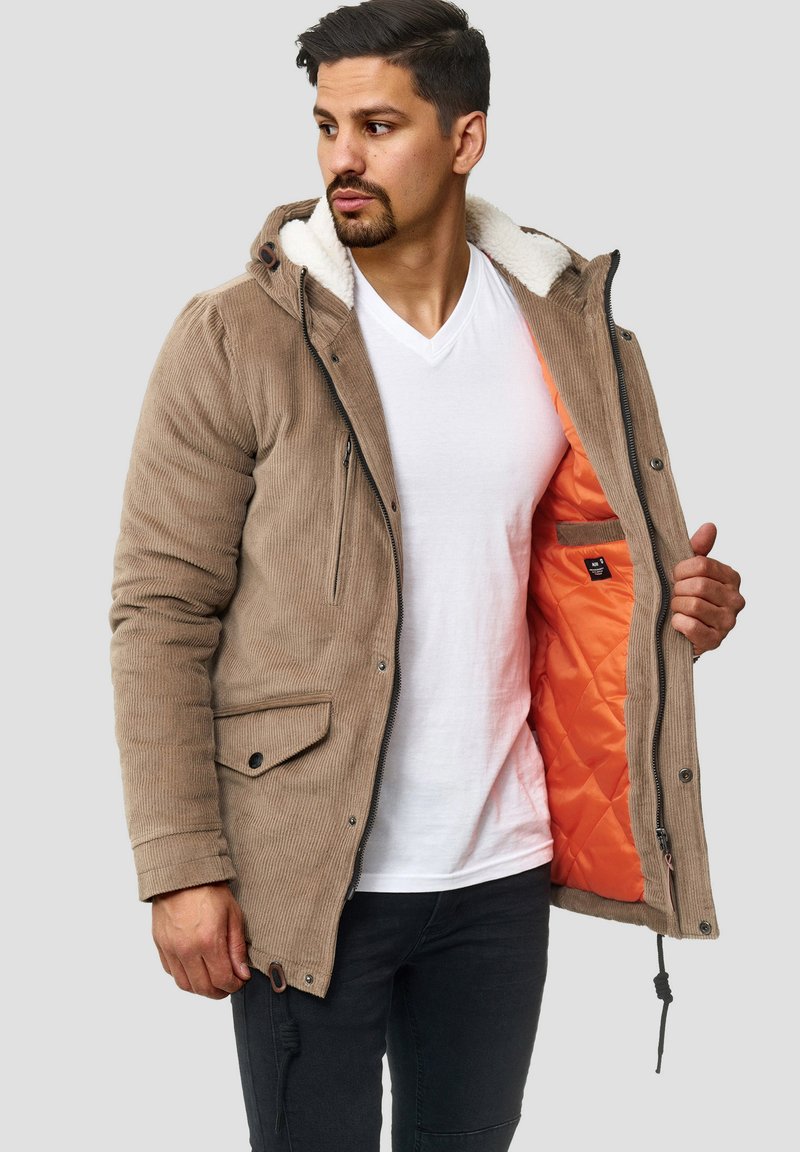 Mand iført en brun fløjlskjorte med hvid fleece-foring, der viser en klar orange quilted inderside, klædt i hvid v-hals skjorte og sorte bukser.