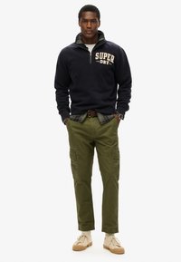 Marine fleece pullover med halvlange lynlås, med et logo i creme; olivenfarvede cargobukser med sidelommer, parret med beige sneakers.