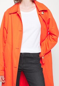 En orange lättviktsjacka med krage, knappstängning och sidofickor, bärs över en vit topp och svarta jeans. Slät textur och skräddarsydd passform.