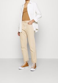Jeans de mezclilla beige, top de tirantes marrón, camisa oversized blanca y zapatillas marrones. Los jeans son de corte recto; la camisa tiene un bolsillo.