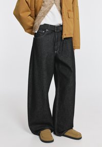 A széles szárú fekete farmernadrág magas derékkal, klasszikus zsebekkel és kontrasztos fehér varrással rendelkezik. Barna dzsekivel és cipővel párosítva.