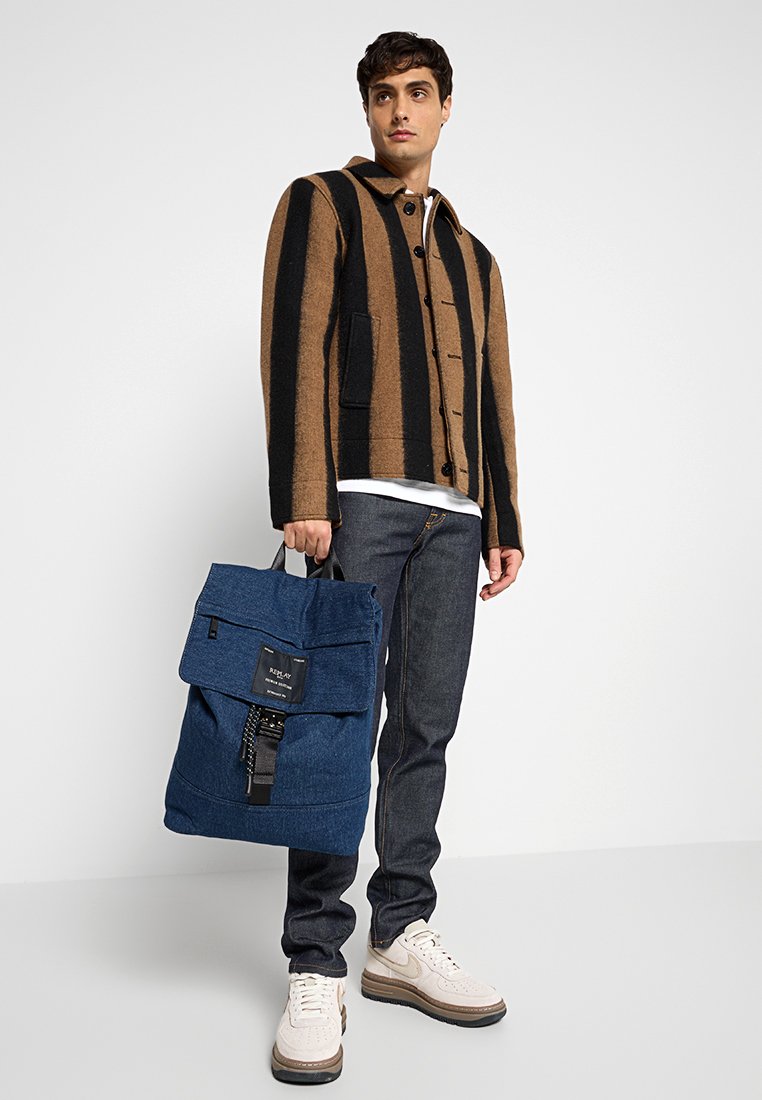 Blauwe denim rugzak met klep en een voorzak met rits, gedragen door een persoon in een gestreepte bruine en zwarte jas en een donkere jeans.