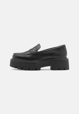 Mocassin en cuir noir avec semelle crantée épaisse et petit talon, présentant une bande décorative sur le dessus, vu de profil sur fond blanc.