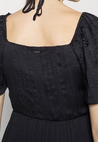 Vestido negro con una tela texturizada, escote cuadrado y mangas abullonadas. Cuenta con un cierre en la parte trasera con lazo y una pequeña etiqueta de marca cerca del cuello.