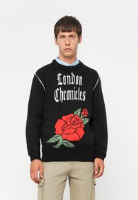 Jonge man in een zwart sweatshirt met de tekst "London Chronicles" en een grote rode rozenafbeelding, gecombineerd met beige cargobroeken en een blauwe kraag die zichtbaar is.