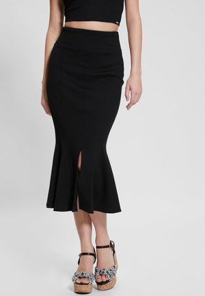 Kobieta nosząca czarną spódnicę midi z wysokim stanem, z przednim rozcięciem i rozszerzanym dołem, w połączeniu z czarno-białymi sandałami na platformie.