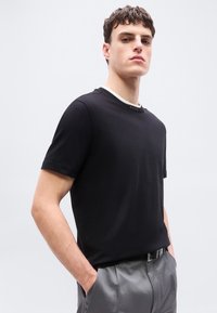 Jeune homme portant une chemise noire à manches courtes et un pantalon gris, debout les mains dans les poches devant un fond clair uni.