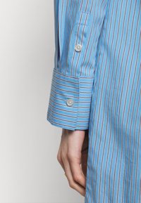 Main reposant sur le côté portant une chemise à manches longues rayée bleue avec poignet boutonné et patte de manche.