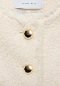 Cremefarbenes, strukturiertes Gewebe mit einem flauschigen Muster. Verfügt über zwei goldene Knöpfe als Akzent. Das Etikett gibt die Größen in EUR, USA und MEX an.