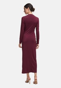 Femme avec les cheveux en chignon portant une robe longue à manches avec une finition maroon et une texture discrète à pois, ainsi que des sandales à talons hauts nude, de dos.