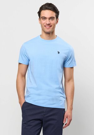 Mladý muž s krátkymi tmavými vlasmi, oblečený v svetlomodré tričko s krátkym rukávom a tmavomodré nohavice, usmievajúci sa s jednou rukou v vrecku.