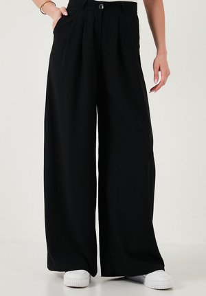 Isik, kes kannab kõrge vöökohaga musta laiade säärtega pükse, millel on voldid ja küljetaskud, ning valgeid tossusid, seisab ühevärvilise tausta ees.