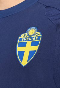 Έμβλημα της εθνικής ποδοσφαιρικής ομάδας της Σουηδίας με κίτρινο σταυρό σε μπλε ασπίδα και κείμενο «SVERIGE» σε σκούρο μπλε αθλητικό φανέλα.