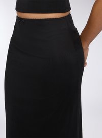 Gonna nera, aderente, realizzata in un tessuto liscio. Presenta un orlo a zigzag discreto e un piccolo dettaglio a fiocco in vita. Termina appena sopra la caviglia.