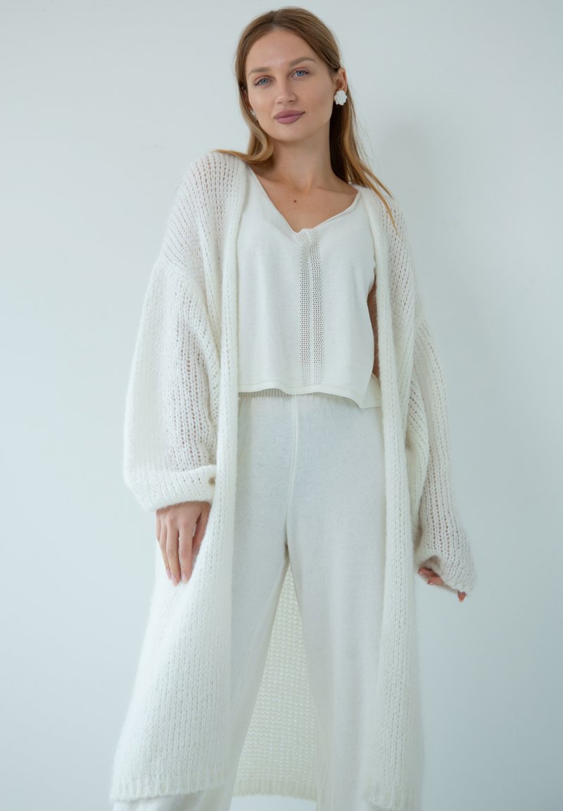 Biała dziergana kardiganka z długim rękawem, z teksturowanym wzorem, zestawiona z białą bluzką z krótkim rękawem i białymi dopasowanymi spodniami.