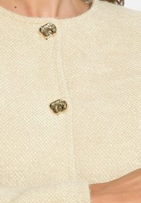 Bežový strukturovaný kardigan s dvěma lesklými zlatými knoflíky na límci, zdůrazňující jednoduchý design a jemnou látku.