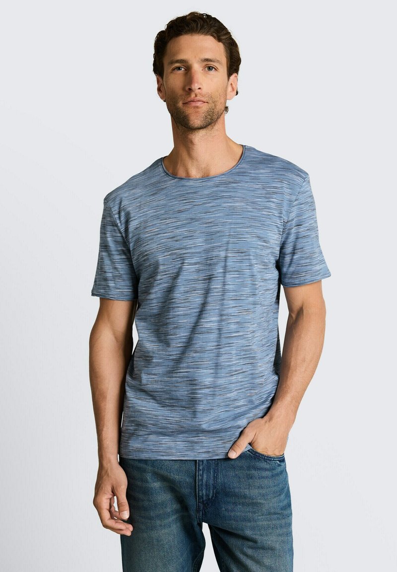 Mann mit kurzen lockigen Haaren, der ein blau gemustertes kurzärmeliges T-Shirt und eine blaue Jeans trägt, eine Hand in der Tasche, neutraler Hintergrund.