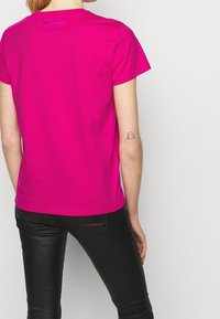 Mujer con camiseta de manga corta de color rosa brillante y pantalones de cuero negros, mostrando un pequeño tatuaje delineado en su antebrazo izquierdo.