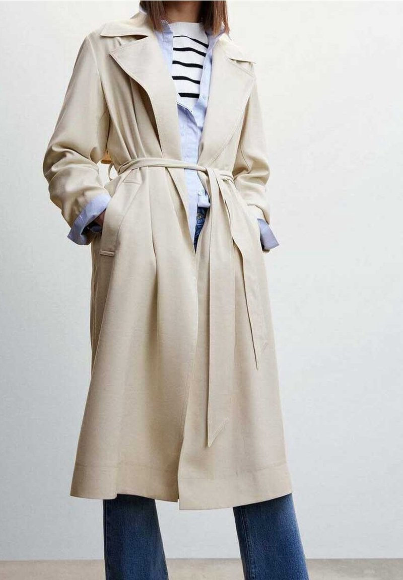 Femme portant un trench coat beige ceinturé par-dessus une chemise rayée et un jean bleu, les mains dans les poches du manteau.