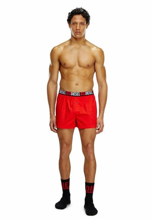Rote Boxershorts mit einem schwarzen, gebrandeten Bund. Modell steht und trägt schwarze Socken mit roter Schrift. Glattes Material, lässige Passform.