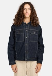 Giacca di jeans di colore blu scuro con due tasche sul petto, chiusura con bottoni e cuciture a contrasto. Presenta un colletto e maniche lunghe.