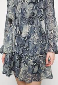 Šaty s hadím vzorem v modré a béžové barvě, s dlouhými rukávy, nařaseným pasem a vrstveným volánkovým lemem. Průsvitná textura s tekutým designem.