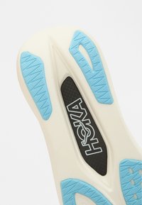 Λευκή σόλα αθλητικού παπουτσιού με υφασμένα μπλε λάστιχα, μια μαύρη περιοχή με λογότυπο και ξεχωριστό σχέδιο για πρόσφυση και στήριξη.