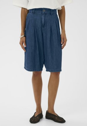 Laza szabású sötét farmer rövidnadrág elülső redőkkel, fehér inggel és fekete szövött slip-on cipővel, fehér háttéren.