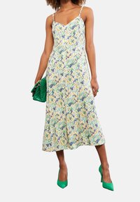 Blumen-Spaghetti-Trägerkleid in Hellblau, Gelb und Grün. Mit einem taillierten Oberteil und einem fließenden Midikleid. Kombiniert mit einer grünen Handtasche.