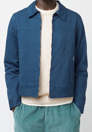 Chaqueta azul marino con un corte entallado, cierre delantero y detalles de costura sutiles. Combinada con un suéter crema y pantalones de pana verde azulado.
