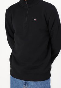 Sudadera de forro polar negra con media cremallera y cuello alto, cremallera frontal y un pequeño parche con logo en el pecho. Textura suave y diseño ajustado.