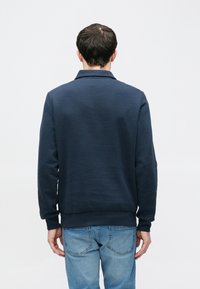 Tumesinine puuvillasegusest dressipluus, millel on krae, soonikkoes mansetid, sile tekstuur ja mugav lõige. Sobitatud siniste teksapükstega.