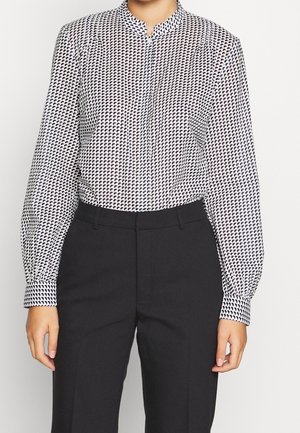 Nainen, joka pukeutunut mustavalkoiseen kuvioituun pitkähihaiseen puseroon, joka on sullottu korkeavyötäröisiin mustiin housuihin, seisoo rentoihin käsiin.