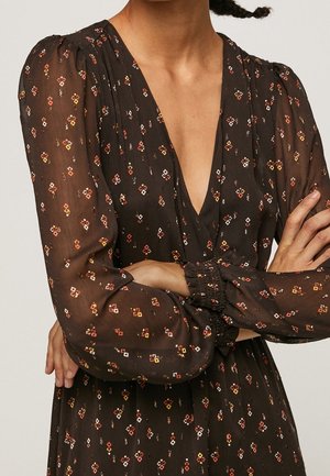 Femme portant une robe sombre semi-transparente avec des motifs géométriques rouges, jaunes et blancs, des longues manches bouffantes et un décolleté en V profond, les bras croisés.