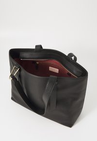 Black leather tote bag featuring a textured exterior, dual top handles, and a red interior with a zippered pocket and branding detail.