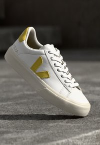 Sapatilha de couro branca com detalhes em amarelo, apresentando uma sola plana, bico redondo e seis furos para atacadores. Design minimalista com costuras visíveis.