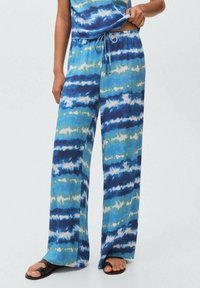Pantaloni largi bleu tie-dye cu talie elastică, purtați cu un top asortat și sandale negre cu vârf deschis, pe un fundal deschis.