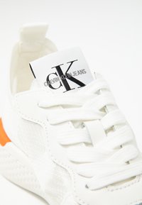 White sneaker with textured fabric, white laces, an orange accent on the heel, and a Calvin Klein Jeans logo on the tongue label.