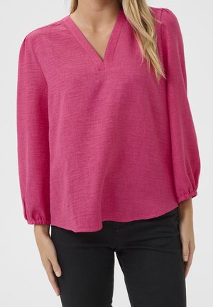 Woman wearing a bright pink long-sleeve blouse with a V-neck and black pants, standing against a plain background.