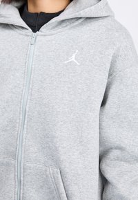 Persona che indossa una felpa con cappuccio e zip color grigio chiaro, con un piccolo logo bianco Jumpman sul petto a sinistra.