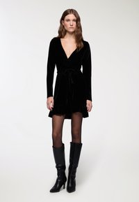 Czarna welurowa sukienka kopertowa z długimi rękawami, głębokim dekoltem w kształcie litery V, przewiązywanym w pasie oraz falbanianym dołem. Noszona z wysokimi na kolana czarnymi skórzanymi botkami i rajstopami w kropki.
