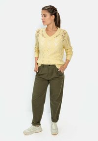 Helles gelbes Strickpullover mit V-Ausschnitt und offener Spitzenoptik, kombiniert mit locker sitzenden olivgrünen Hosen und hellbeigen Sneakers.