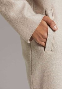 Beige Wollmantel aus strukturiertem, weichem Material mit einer Seitentasche. DieÄrmel haben einen klaren, geraden Schnitt mit minimalen Nähten.