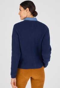 Maglione lavorato a maglia blu navy con texture a coste e scollo rotondo, abbinato a un colletto in denim azzurro. Indossato con pantaloni beige.